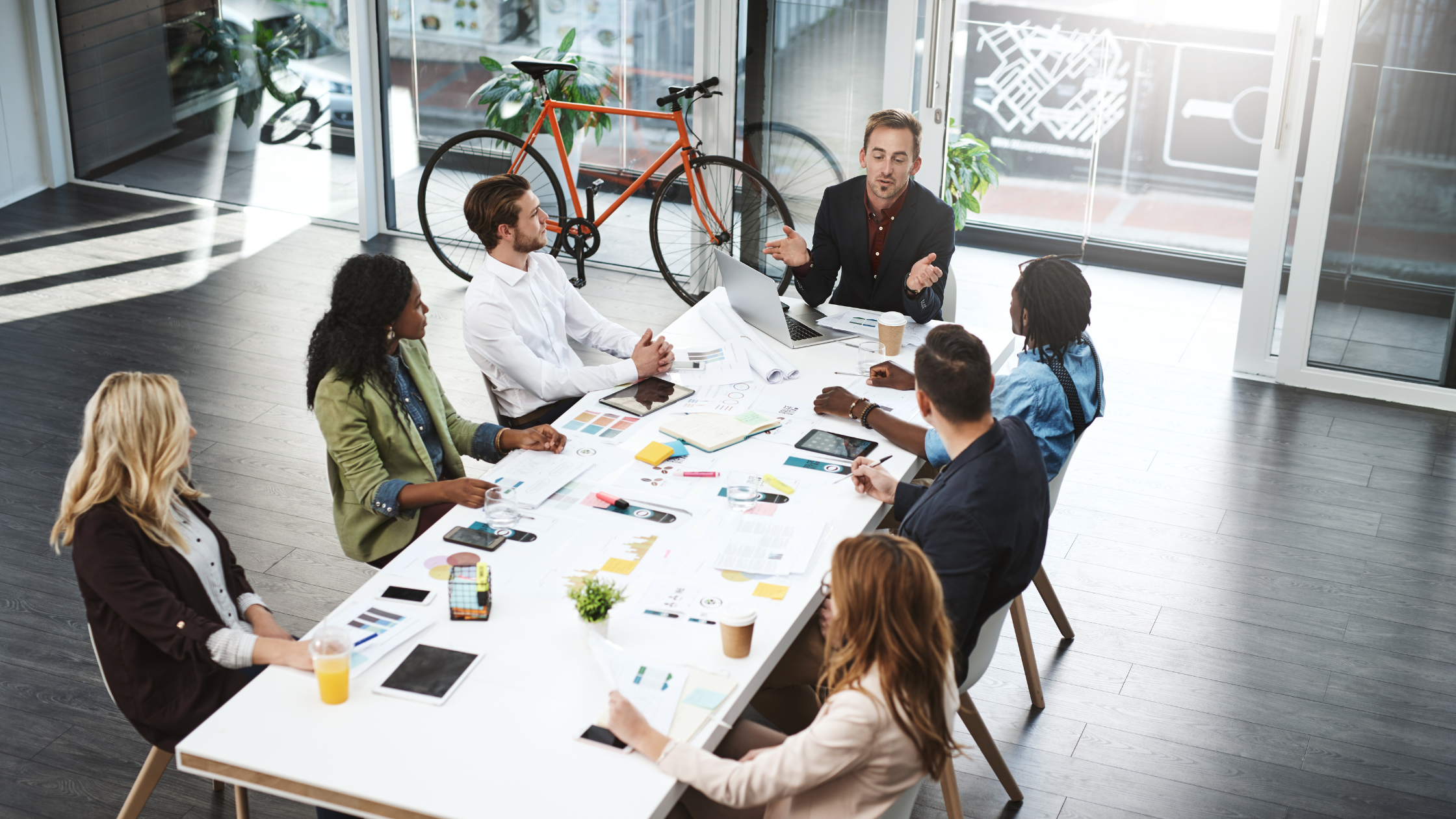 A group of seven professionals sitting around a table in the workplace, having a meeting.